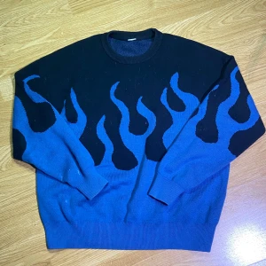 Blå och svart tröja med flam-mönster - Cool stickad tröja i blått och svart med eldflammor som motiv. Oversized passform och rund halsringning. Perfekt för dig som vill sticka ut med en unik stil.