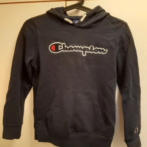 Mörkblå hoodie från Champion, strlk 138/142 - Mörkblå hoodie från Champion med broderad logga på bröstet och ena ärmen. Storlek 138/142 (barn Medium).