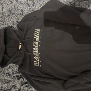 Svart hoodie från Napapijri - Säljer en svart hoodie från Napapijri med stor logga framtill. Tröjan har huva med dragsko och en klassisk, avslappnad passform. Perfekt för dig som gillar streetwear och vill ha något bekvämt och stilrent.