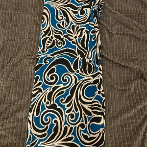 Blå och svart mönstrad kjol - Säljer en kjol med blå, svart och vit swirl-mönster. Kjolen har omlottknytning i midjan och är tillverkad i ett lätt och luftigt material, perfekt för varmare dagar. Snygg och iögonfallande design som sticker ut.