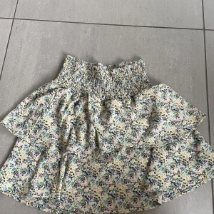 Blommig volangkjol med smock i midjan - Supersöt kort kjol med blommigt mönster i pastellfärger och volangdetaljer. Kjolen har smockad midja som ger en bekväm passform och är perfekt för varma dagar.