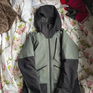 Grön och svart skaljacka från Everest  - Säljer en grön och svart skaljacka från Everest med huva och dragkedjor framtill. Jackan har flera praktiska fickor med dragkedja och en sportig look. Perfekt för utomhusaktiviteter när vädret är ostadigt för att den håller dig varm och den är också vattentät. Det ända felet är att den är riven på ett ställe men det är inte stort