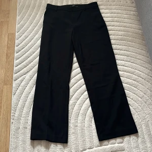 Svarta kostymbyxor från Gina Tricot - Klassiska svarta kostymbyxor från Gina Tricot med raka ben och normal passform. Byxorna har hällor för bälte, diskreta sidfickor och stängs med dragkedja och knapp. Perfekta till en stilren look.