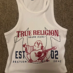 Vit linnetopp från True Religion - Vit croppad linnetopp från True Religion med tryck i rött och blått framtill. Motiv av en Buddha som spelar gitarr och texten 'True Religion Brand Jeans'. Perfekt för en avslappnad stil.