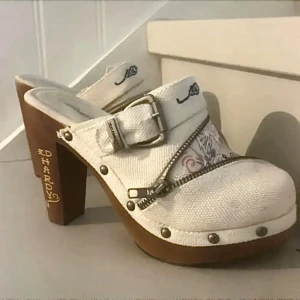 Ed Hardy klackskor - Säljer ett par unika Ed Hardy klaackar med hög klack och coolt koinarp-mönster. Skorna är vita med detaljer i form av dragkedja och spänne. Perfekta för en cool stil!