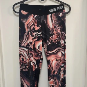 Nike Pro mönstrade träningstights - Nike Pro tights med unikt abstrakt mönster i svart, rosa och vitt. Tightsen har bred resår i midjan med Nike Pro-logga och är perfekta för träning. Sitter tight och följer kroppen för maximal rörelsefrihet.