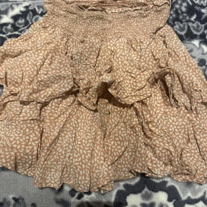 Beige volangkjol med mönster - Supersöt beige kortkjol med vitt bladmönster och flera volanger. Kjolen har smock i midjan för en bekväm passform och är perfekt för varma dagar.