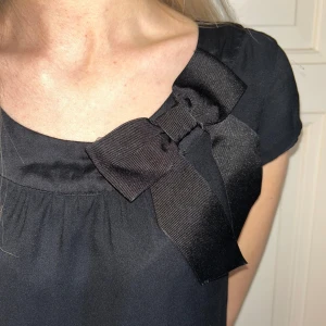 Svart finare klänning med rosett från H&M - kortklänning - En stilren, lite finare svart klänning från H&M med korta ärmar och en stor dekorativ rosett vid halsringningen. Klänningen har en rak passform och är tillverkad i ett mjukt, slätt material som faller snyggt. Ser oanvänd ut. 