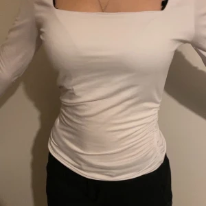 Vit långärmad topp med rynkade sidor - Säljer en vit långärmad topp med fyrkantig ringning och snygga rynkade detaljer längs sidorna. Toppen har en tight passform och är tillverkad i ett stretchigt material som sitter bekvämt på kroppen.