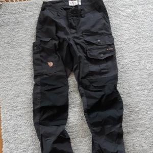 Svarta byxor Vidda Pro W Fjällräven G-1000 - Svarta byxor från Fjällräven i slitstarkt G-1000-material. Byxorna har flera stora fickor med knappar på benen och klassisk Fjällräven-logga. Perfekta för uteliv och äventyr. Regular fit Mid waist. Ganska höga i midjan, sköna vandringsbyxor.