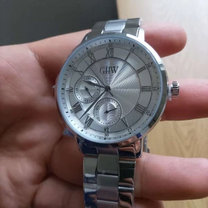 Silverfärgad klocka från GHW - Stilren klocka från GHW i helsilver med romerska siffror på urtavlan och två mindre urtavlor. Armband och boett i metall med polerad finish. Passar dig som gillar klassisk design och detaljer. Klockan är heller ej använd, fick ärva den av en släkting tillsammans med andra klockor. Plasten på urtavlan är kvar och batteri tillkommer såklart Nypris på klockan ligger på strax över 1300kr