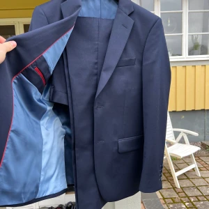 Mörkblå kostymkavaj med ljusblått foder - Mörkblå kostym med matchande byxor och kavaj. Kavajen har en ljusblå innerfoder med röda detaljer och en bröstficka samt två fickor på framsidan. Byxorna är klassiskt skurna och kompletterar kavajen väl. Perfekt till balen.  Jacken är av storleken 44 short och byxorna 31/30 både av passform slim fit  Jag som har på mig kostymen i bild 4 står i mitten och var cirka 165cm lång  Endast använd 2 gånger och i nyskick  Nypris cirka 2000kr