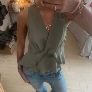 Olivgrön omlottopp med volang - Snygg olivgrön omlottopp med volangdetalj nertill. Ärmlös modell med v-ringning, perfekt till jeans eller kjol. ÄLSKAR