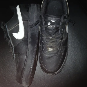 Svarta Nike Air Force 1 sneakers - Säljer ett par klassiska svarta Nike Air Force 1 sneakers med vit swoosh. Skorna har rund tå, platt sula och snörning framtill. Perfekta för en streetwear-look och passar till det mesta. Ikonisk design med Nike-logga på sidan. 8/10 skick lite dammiga för dem har inte används på länge små crease som går att få bort dammet får du bort enkelt, små skador på swooshet men märks inte och baksidan är lite creasade