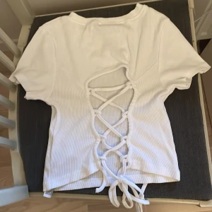 Vit ribbad topp med snörning från ett av NewYorkers märken  FBSISTER - Säljer en vit ribbad topp från ett av NewYorkers märken FBSISTER i storlek XS. Toppen har korta ärmar och snygg snörningsdetalj i ryggen som ger en unik look. Perfekt för dig som vill ha något stilrent men ändå speciellt.