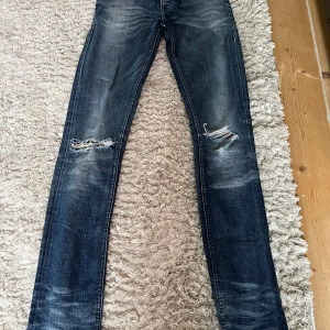 Marineblå Nudie Jeans - Säljer ett par Marineblå jeans från Nudie Jeans med snygga slitningar på båda benen. Klassisk femficksmodell med dragkedja och knapp. Jeansen har en slim fit passform och är tillverkade i denim. Jeansen är W28/L34 men passar mer W28/L32. Dom är perfekta för en stilren look.