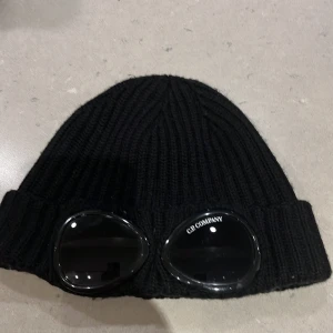 Svart ribbstickad mössa med goggles från C.P. Company - Svart ribbstickad mössa från C.P. Company med inbyggda goggles framtill. Klassisk modell med uppvikt kant och tydlig logga på ena glaset. Perfekt accessoar för en unik och sportig look.