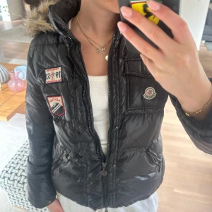 Svart dunjacka med pälsdetalj från Moncler - Säljer en svart dunjacka från Moncler med pälsdetalj på huvan och coola patches framtill. Jackan har dragkedja, fickor och stor moncler logga på ryggen. Luvan är avtagbar 