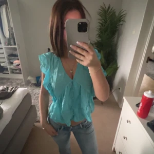 Turkos volangtopp - Säljer en turkos topp med volangdetaljer och v-ringning. Toppen har korta ärmar och är tillverkad i ett lätt material, perfekt för varmare dagar. Snygg att matcha med jeans eller kjol.
