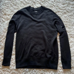 Svart sweatshirt från C.P. Company - Säljer en svart sweatshirt från C.P. Company med deras ikoniska linsdetalj på ärmen. Tröjan har rund halsringning, långa ärmar och är tillverkad i mjuk bomull. Perfekt för en stilren och avslappnad look.