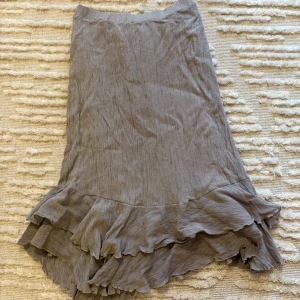 Grå kjol med volang - Säljer en asymmetrisk grå/beige kjol med snygga volangdetaljer längst ner. Kjolen har en mjuk och flowy känsla med lätt struktur i tyget och är längre baktill. Perfekt för dig som gillar en feminin och trendig stil.