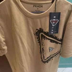 Beige t-shirt från Prada med grafiskt tryck - Säljer en beige t-shirt från Prada i regular fit med ett svart grafiskt tryck på bröstet. T-shirten har rund halsringning och korta ärmar. Perfekt för dig som gillar stilrena plagg med en twist.