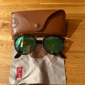 Ray-Ban runda solglasögon brun/grön - Säljer ett par Ray-Ban RB4256 solglasögon med runda bågar i brunsköldpaddsmönster och spegelglas i grön ton. Snygg retrostil med metallbrygga och klassisk Ray-Ban-logga på skalmen. Kommer med originalfodral i brunt och putsduk.