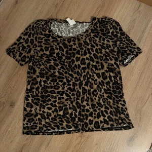 Leopardmönstrad topp från H&M - Säljer en kortärmad topp från H&M med snyggt leopardmönster i bruna och svarta toner. Modellen har rund halsringning och lätt puffade axlar för en trendig look. Tunt och mjukt material som känns skönt mot huden.