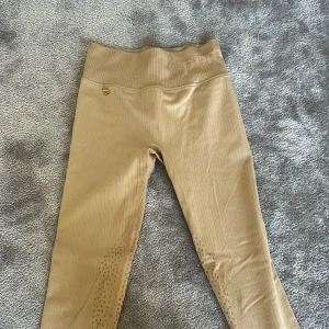 Beige höga ridleggings från Pomme - Snygga beige ridleggings från Pomme med hög midja och ribbad struktur. De har silikonprickar på insidan av benen för extra grepp och är tillverkade i ett stretchigt syntetmaterial. Perfekta för dig som vill ha både stil och funktion i stallet.