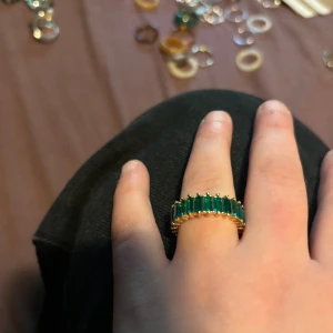 Guldfärgad ring med gröna stenar - Snygg guldfärgad ring med rektangulära gröna stenar runt hela bandet. Ringen har en chunky känsla och de smaragdgröna stenarna ger en lyxig och trendig vibe. Perfekt statement-piece för dig som vill sticka ut.