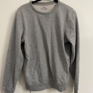 Grå sweatshirt från Gildan  - Enkel och stilren grå sweatshirt från Gildan med rund halsringning och ribbade muddar vid ärmslut och nederkant. Perfekt för en chill och avslappnad look. Tillverkad i mjuk bomullsmix som känns skön mot huden.