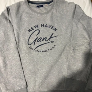 Grå sweatshirt från Gant - Snygg grå sweatshirt från Gant med blå tryck framtill där det står 'New Haven Gant Est. 1949 NHCT U.S.A.'. Tröjan har rund halsringning, långa ärmar och ribbade muddar vid ärmslut och nederkant. Perfekt för en chill och stilren look.