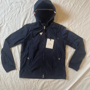 Mörkblå vindjacka från Moncler - Snygg mörkblå vindjacka från Moncler med huva och dragkedja framtill. Jackan har Moncler-logga på ärmen, två fickor med tryckknappar och coola ränder på huvan. Perfekt för blåsiga dagar och enkel att matcha med allt. Materialet är lätt och smidigt syntet. Den är helt ny och aldrig använd mycket bra kvalité och skick.