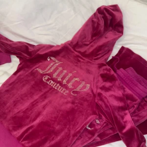 Mörkrosa Juicy Couture hoodie och byxor i velour - Säljer en ikonisk mörkrosa Juicy Couture hoodie i mjuk velour med glittrande strasstenar på ryggen och JC-logga framtill. Tröjan har huva, dragkedja och ribbade muddar. Perfekt för dig som vill ha den klassiska Y2K-looken!