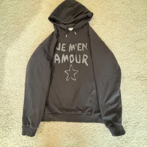 Svart hoodie Gina Tricot med glittertext - Svart hoodie från Gina Tricot med huva och långa ärmar. Framsidan har glittrig text 'JE M'EN AMOUR' och en stjärna. Mjuk insida och ribbade muddar. Perfekt för dig som gillar statement-plagg med lite extra attityd.