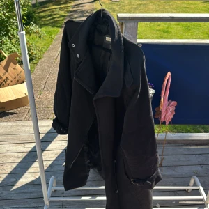 Svart lång trenchcoat från Mango  - Säljer en svart trenchcoat från Vero Moda med snyggt bälte vid ärmsluten och dragkedja framtill. Jackan har en klassisk krage och långa ärmar, perfekt för dig som gillar stilrena och tidlösa plagg. Passar dig som vill ha en clean look i höst och vinter.