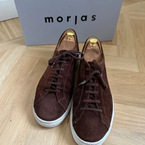 Bruna mockasneakers från Morjas - Snygga bruna sneakers från Morjas i mjuk mocka med vita sulor och klassisk snörning. Låg modell med rund tå och stilren design som passar till det mesta. Perfekta för dig som gillar en clean och tidlös look. Skolåda ingår ej skoblock.