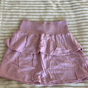 Rosa volangkjol med smock - Supersöt rosa kortkjol från 157 med bred smock i midjan och dubbla volanger. Kjolen är lätt och flowy, perfekt för varma dagar. Den har en ljus pastellrosa ton och är riktigt bekväm att bära.