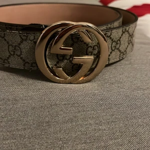 Gucci GG Supreme bälte med guldspänne - Snyggt Gucci GG Supreme bälte med klassiskt monogrammönster i grått och svart. Bältet har ett stort, rundat guldspänne med dubbla G-loggan och är gjort i skinn med canvasdetaljer. Perfekt accessoar för att lyfta din outfit med lyxig känsla.