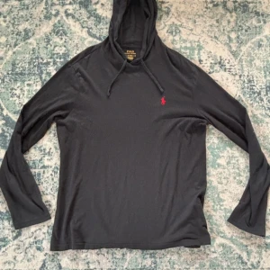 Svart hoodie från Polo Ralph Lauren - Svart hoodie från Polo Ralph Lauren med klassisk röd broderad logga på bröstet. Tröjan har huva med snörning och långa ärmar. Tillverkad i mjuk bomull, perfekt för en chill och stilren look.