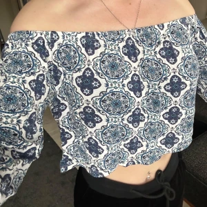 Offshoulder croppad blus med blått mönster - Säljer en superfin offshoulder croppad blus med blått och vitt mönster. Toppen har långa ärmar och resår upptill för att sitta snyggt över axlarna. 