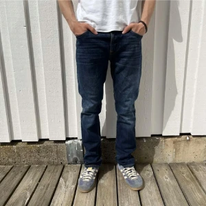 Nudie jeans grim trim  - Snygga mörkblå jeans från Nudie Jeans med klassisk femficksdesign och raka ben. Jeansen har kontrastsömmar och läderpatch bak i midjan. Perfekta för dig som gillar en stilren och tidlös look. Jeans är i riktigt bra skick säljes dock för att de är för små. 