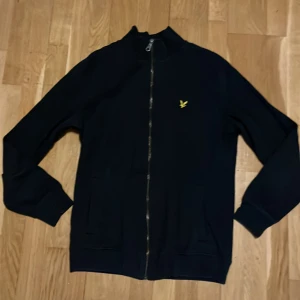 Svart ziptröja från Lyle & Scott - Svart jacka med hel dragkedja från Lyle & Scott. Klassisk design med ribbade muddar och liten gul logga på bröstet. Perfekt för en clean och stilren look. Långärmad och med två fickor framtill.
