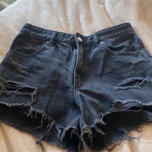 Svarta slitna jeansshorts - Snygga svarta jeansshorts med hög midja och rejält slitna detaljer. Råa kanter och flera hål ger en cool, edgy vibe. Perfekta för sommardagar när du vill sticka ut lite extra. Klassisk femficksmodell med knapp och dragkedja.