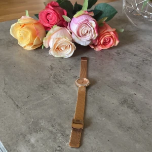 Guldig klocka från Ivy N Rox - Snygg klocka i guld från Ivy N Rox med mesharmband i metall och rund urtavla. Urtavlan är dekorerad med små stenar runt kanten och har minimalistiska markeringar. Perfekt accessoar för att lyfta din outfit.