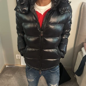 Svart Moncler pufferjacka med huva - Snygg svart pufferjacka från Moncler med glansig finish och huva. Jackan har dragkedja framtill, fickor på sidorna och en Moncler-logga på ärmen. Perfekt för kalla dagar och ger en cool streetstyle-vibe. Pirslap finns nfc finns helt ny 