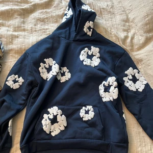 Blå hoodie med vita blommor Denim Tears - Säljer en marinblå hoodie från Denim Tears med stora vita blommönster över hela tröjan. Tröjan har huva, känguruficka och är långärmad. Perfekt för dig som vill sticka ut med en unik och trendig look.