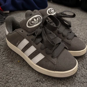 Adidas Campus grå/vita sneakers - Säljer ett par Adidas Campus sneakers i mörkgrå mocka med klassiska vita ränder och vit sula. 😸Skorna har breda gråa snören och Adidas logga på plösen.  Endast använda ett par gånger - så skicket är bra även om det finns ett par tecken på användning 😇Pris kan diskuteras! 