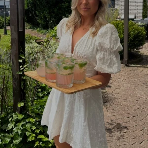 Vit kort klänning med puffärm från Adoore - Superfin vit kort klänning med broderade blommor, djup v-ringning och puffiga korta ärmar. Klänningen har markerad midja, knappar framtill och lätt utsvängd kjol. Perfekt för sommarens alla tillfällen! Använd 2 gånger.