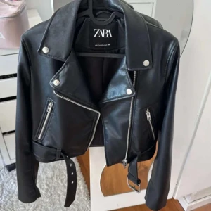 Svart skinnjacka från Zara - Snygg svart skinnjacka från Zara med klassisk bikerstil. Jackan har silvriga dragkedjor, bälte i midjan och breda slag. Perfekt för dig som vill ha en edgy look med coola detaljer.
